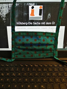 Dieses Farbfoto von Thorsten Hülsberg zeigt einen Laptop mit der Website von hÜlsberg – Die Sache dem Ü und eine davor gehangene Gesichtsmaske.